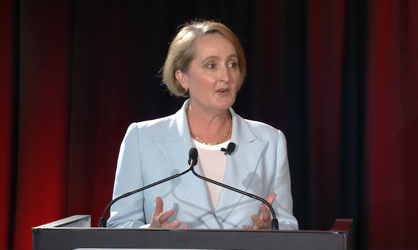 A woman wearing a light blue suit speaks behind a microphoned lecturn with a deep red curtain behind.