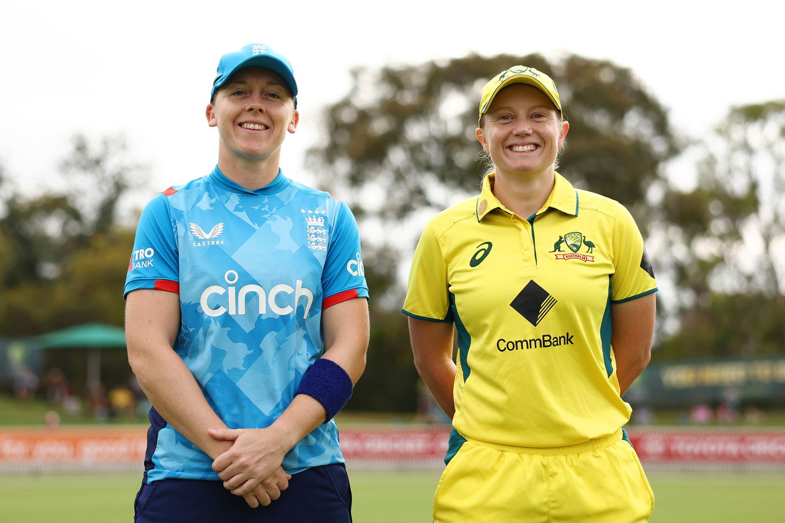 Two women, one in a blue England shirt and cap, and the other in a yellow Australia one, stand smiling and facing the camera.