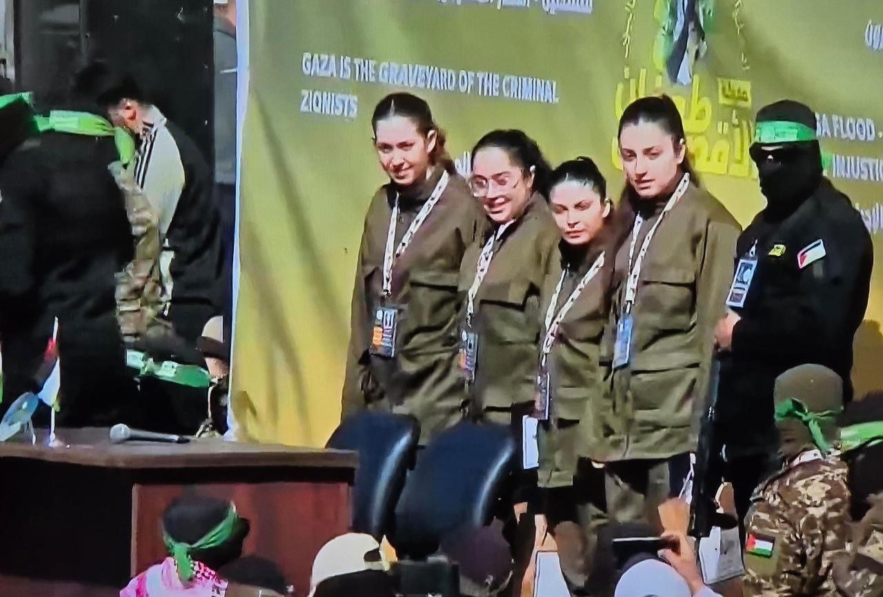 Four women and a masked militant on a stage looking on.