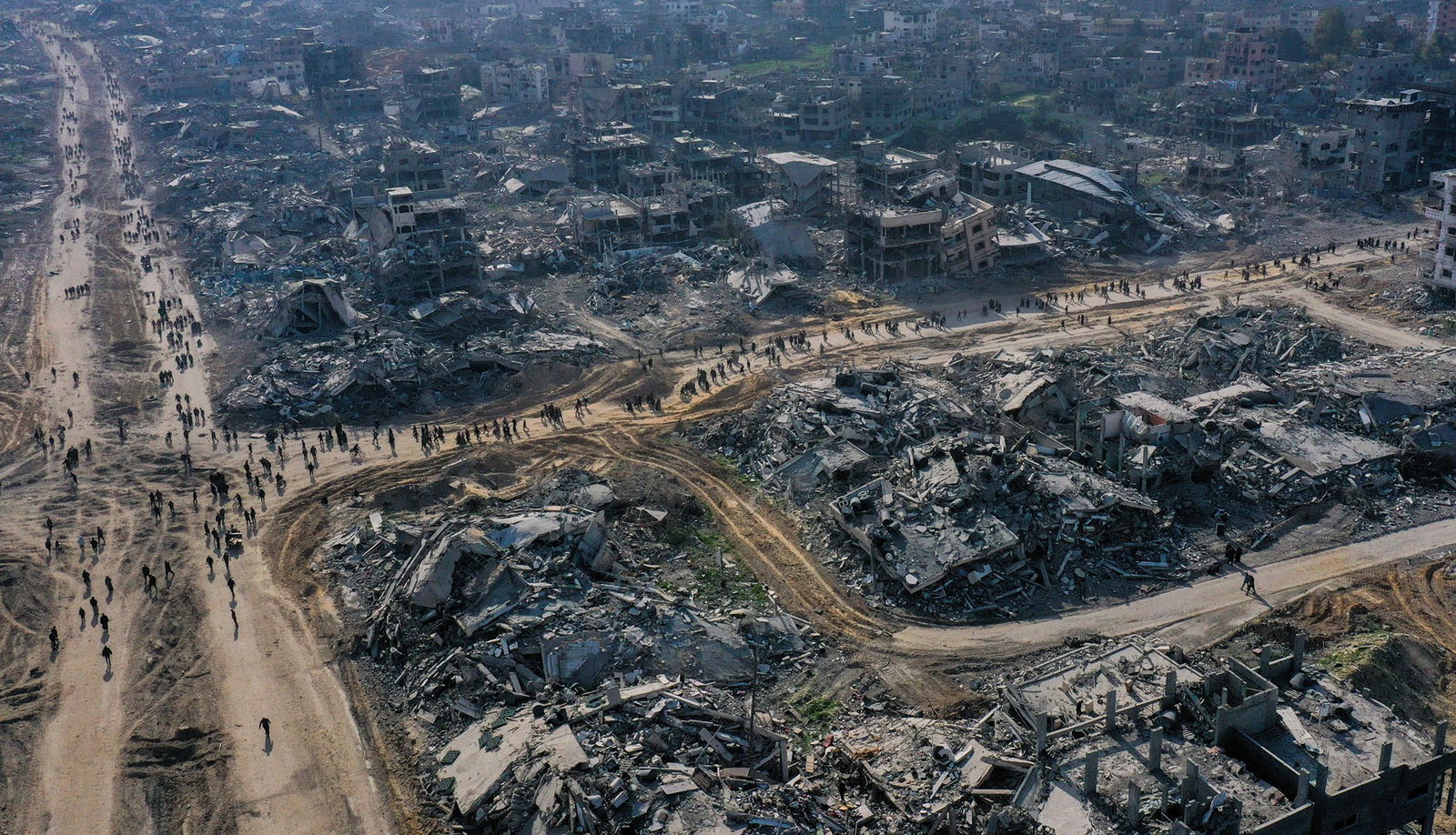 A drone shot showing a neighbourhood of apartment buildings entirely bombed out, with a number of people walking down the road.