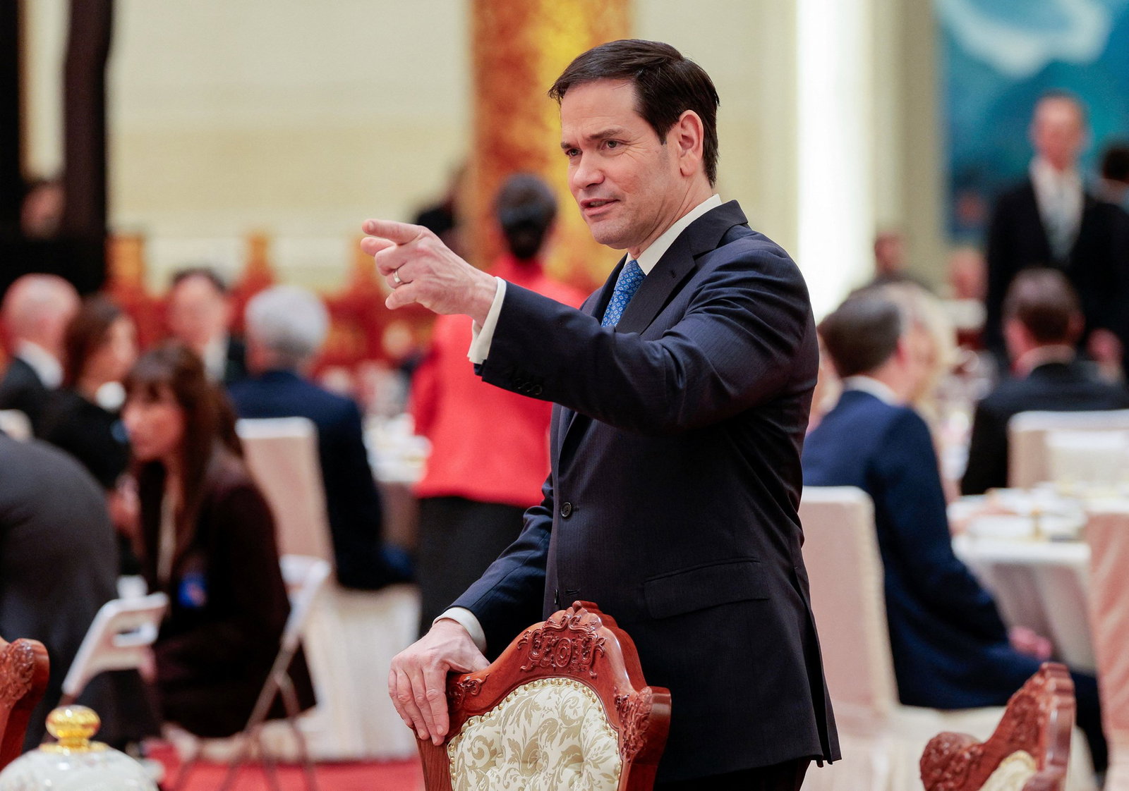 Marco Rubio stands behind an antique dining chair and points at something