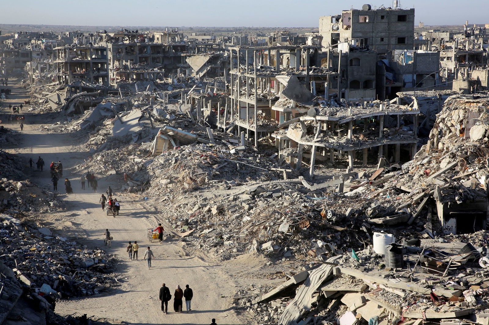 Damaged concrete building lay in ruins as people walk along a dirt road next to them