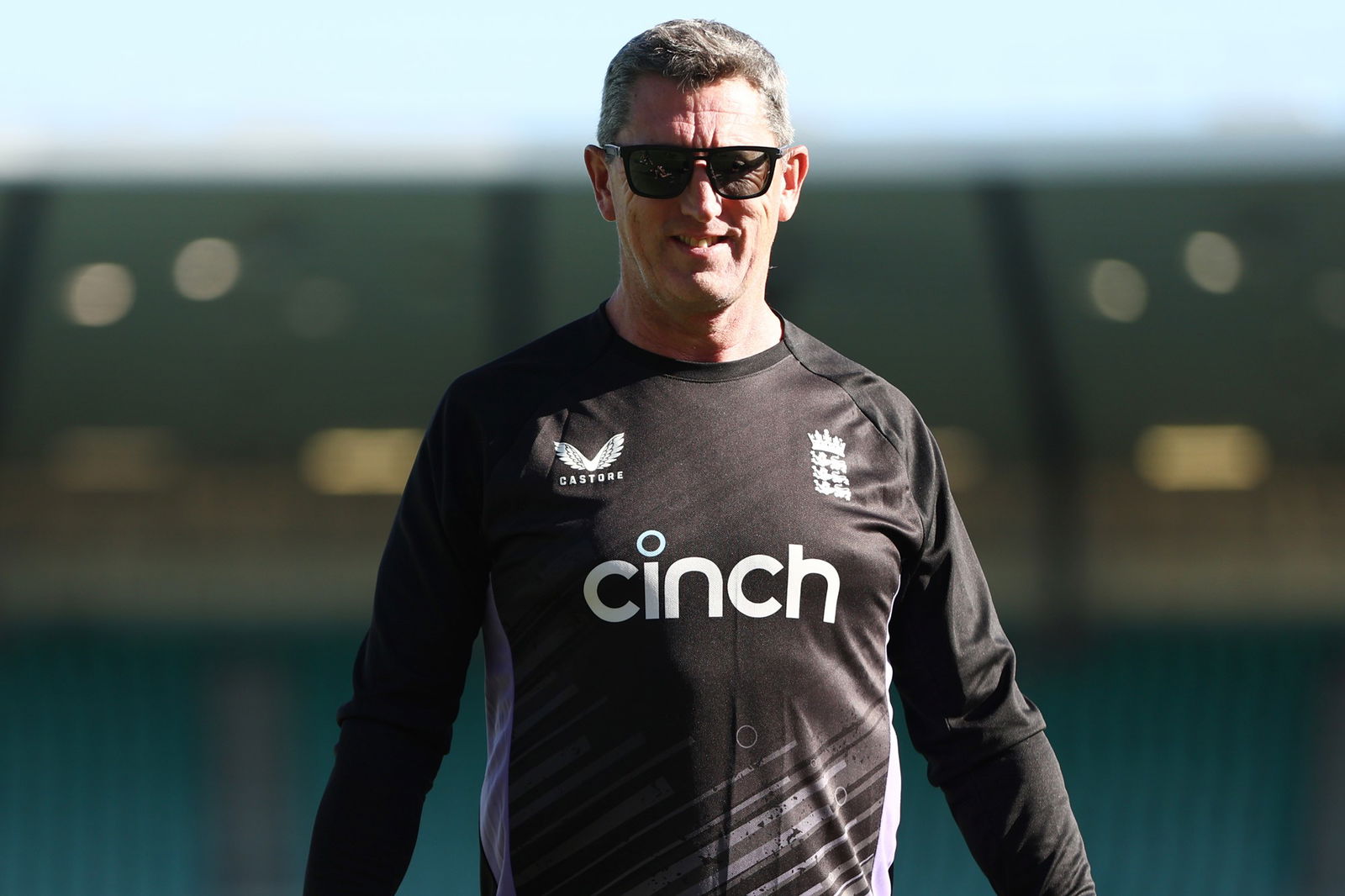 A man wearing a black England cricket training top and sunglasses smiles at the camera