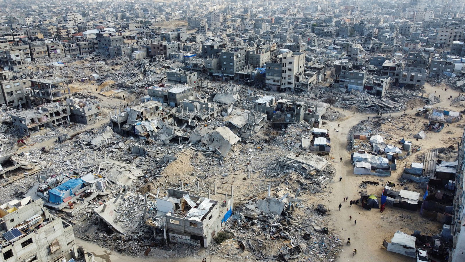 A drone view shows Palestinians walking past the rubble of houses and buildings destroyed in Israeli strikes