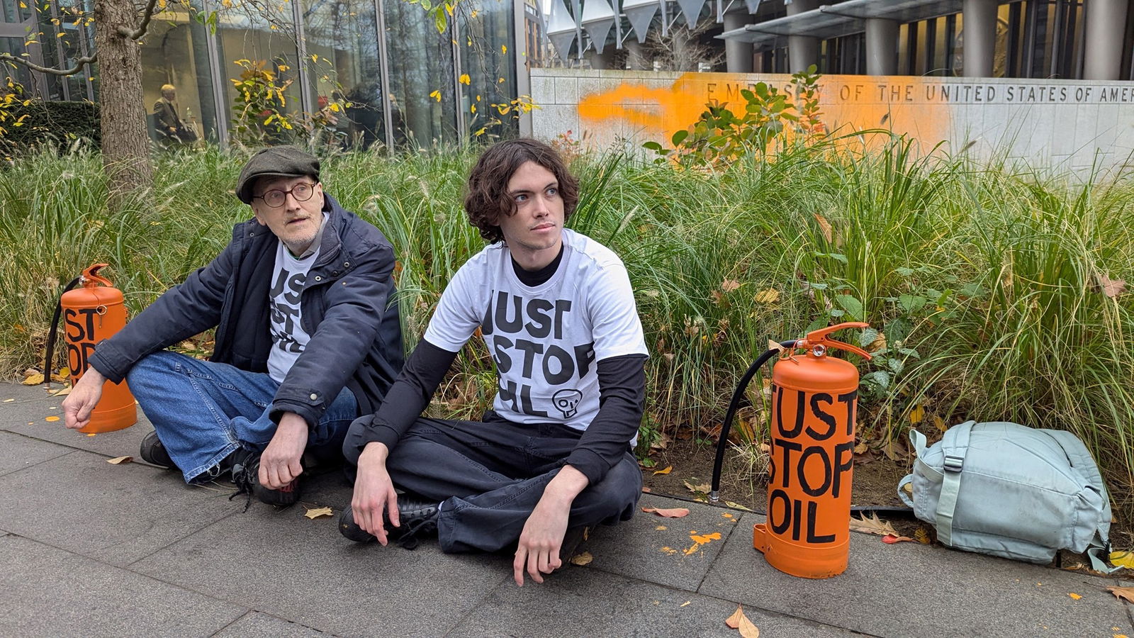 Two people sit in front of a wall painted orange in just stop oil shirts