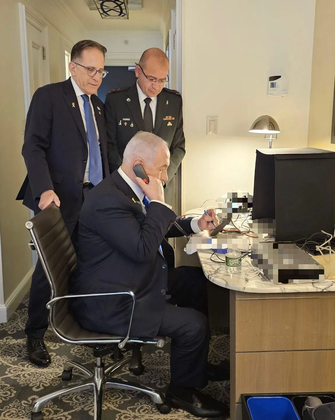 A man sitting at a desk on the phone, while two other men look on.