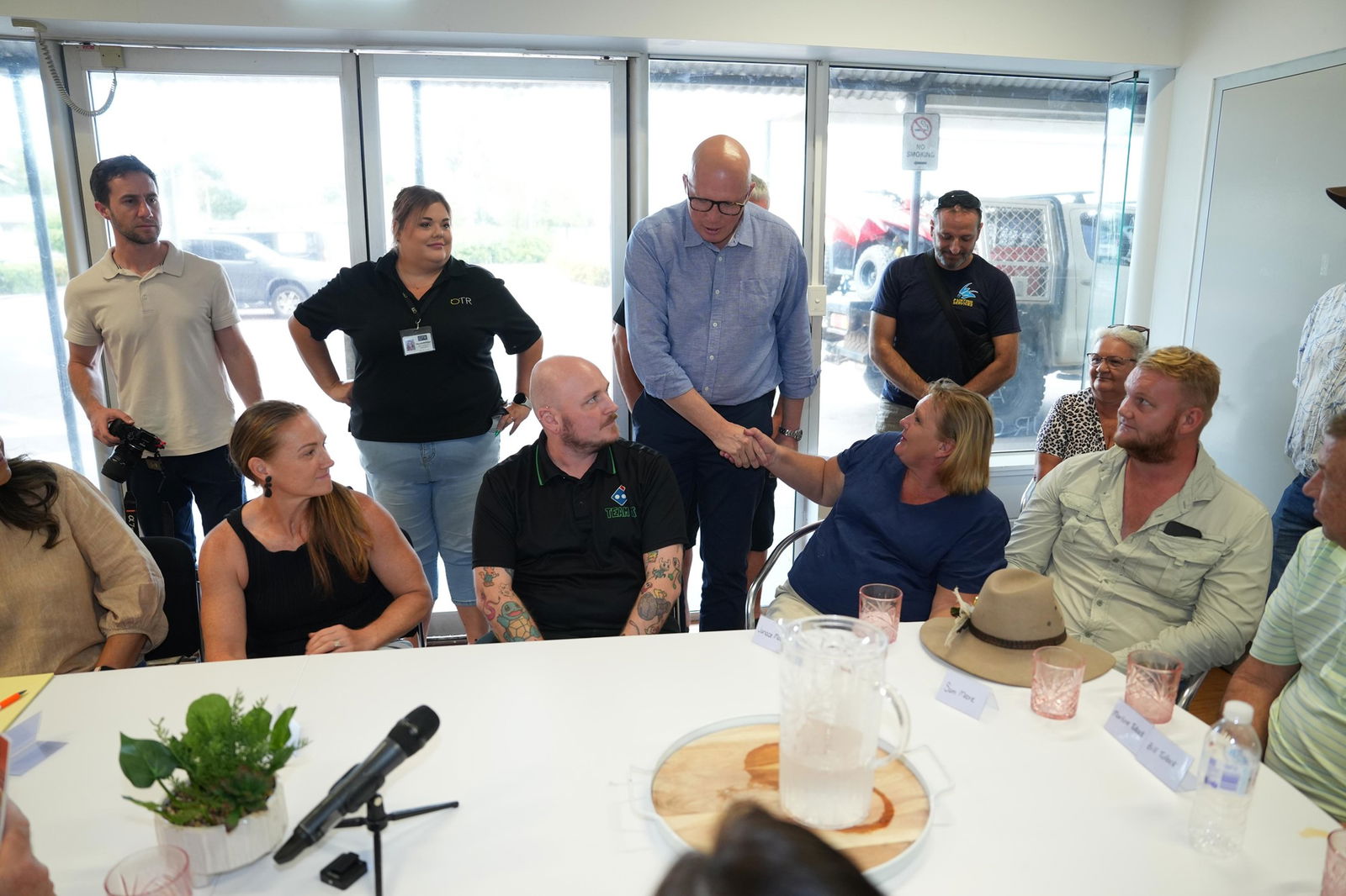 Peter Dutton shakes hands with a woman seated at a big table with a number of other people.