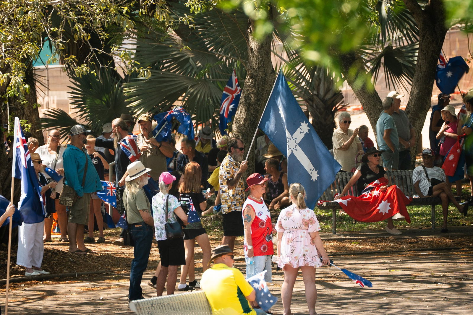 Una multitud de personas paradas en un parque, con algunas sosteniendo banderas australianas. Una persona está sosteniendo una bandera de Eureka y otra tiene una bandera roja de Ensign sobre su regazo.
