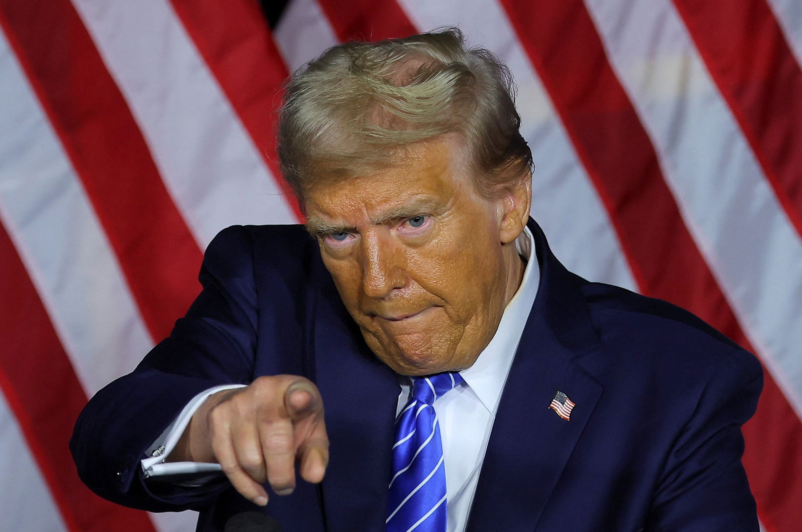 Trump points a finger and purses his lips as he stands in front of an American flag. 