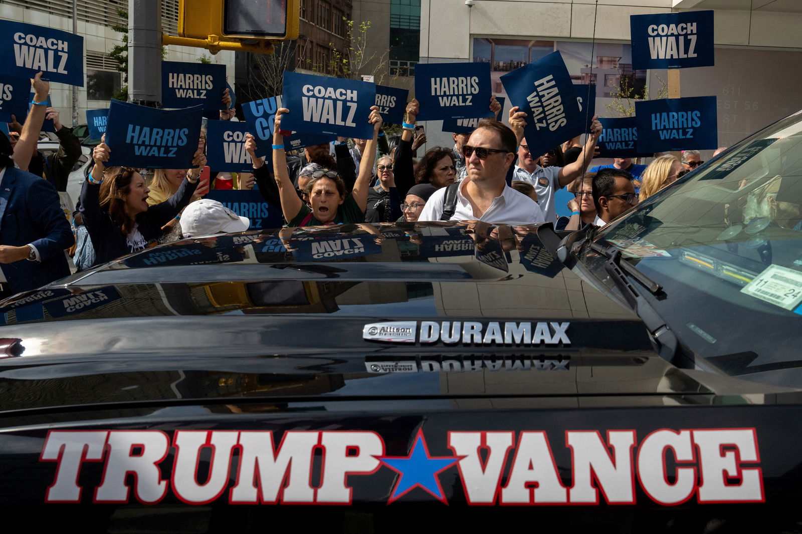 People hold “Harris Walz” and “Coach Walz” signs behind a large black vehicle that says “Trump Vance”