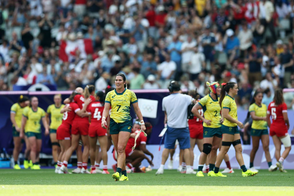 A woman in gold and green walks away from celebrating Canadians in red and her dejected teammates.