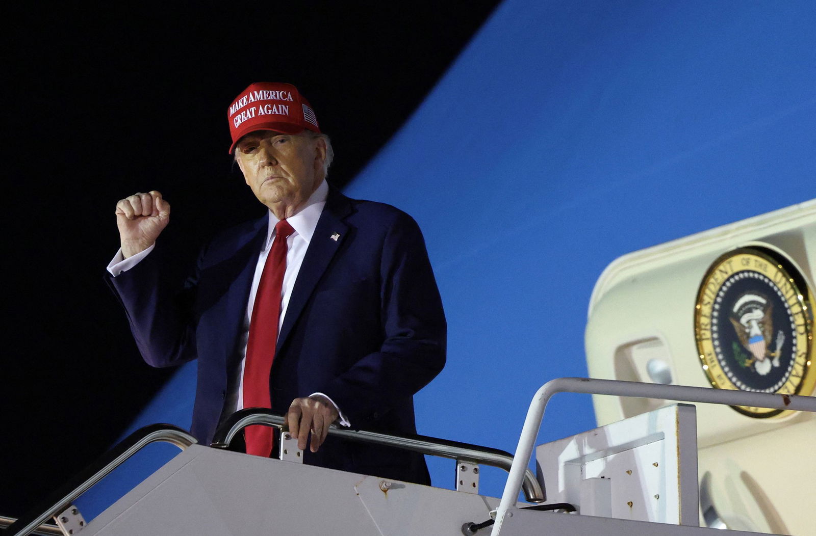 Donald Trump in a blue suit, red tie and red Make America Great Again hat descending an aircraft stairway at night with his left hand on a rail