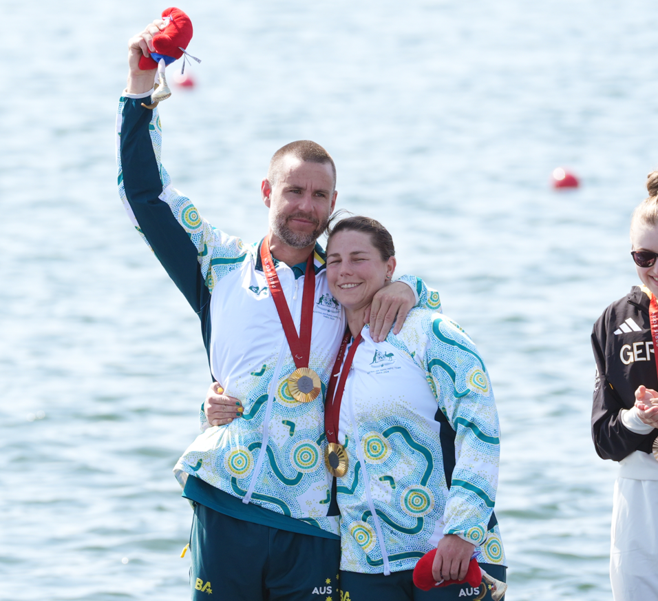 A man and a woman stand in front of blue water wearing gold medals. The man has one hand in the air and the other around the woman. They are both smiling and wearing green and white, Australia branded tracksuit tops.