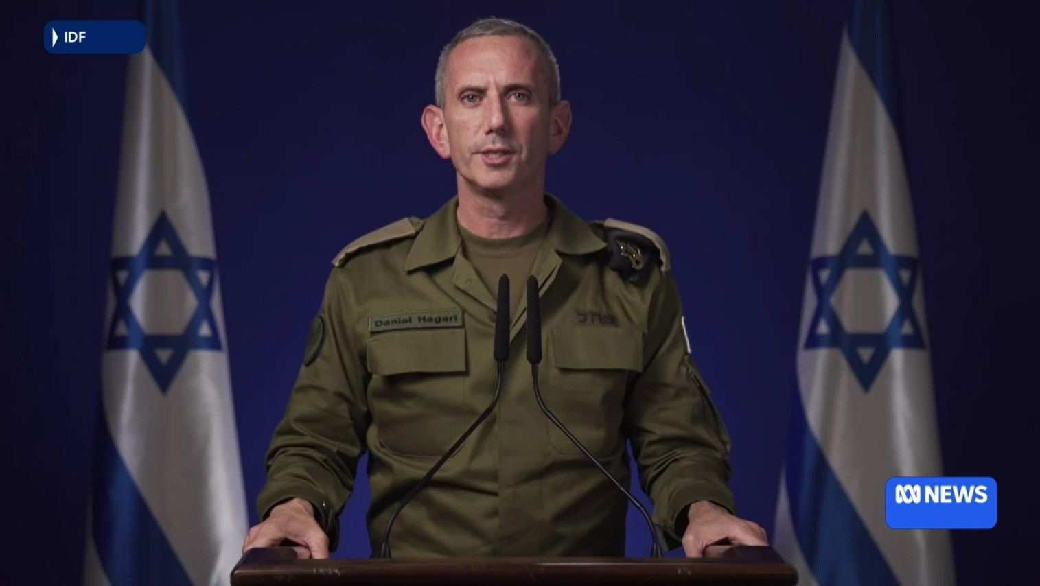 A man in uniform speaking to a camera, with Israeli flags visible behind him.