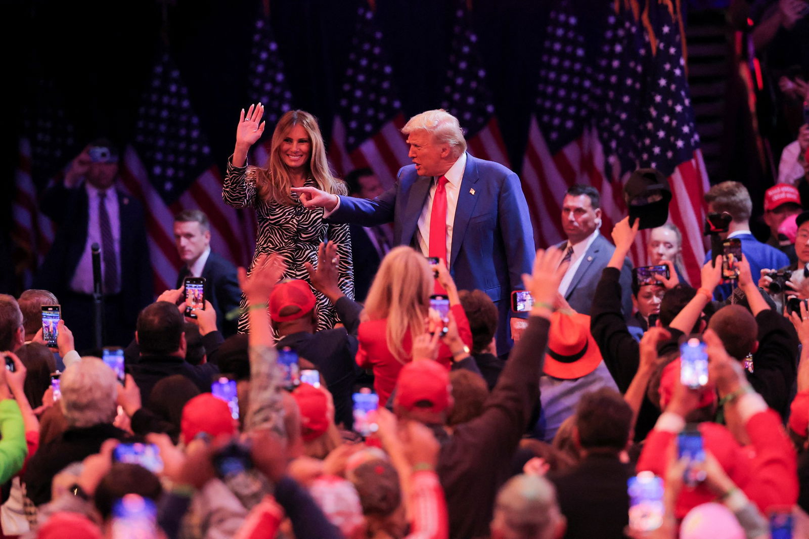 Donald Trump stands on a stage swamped by supporters, pointing at someone in the crowd