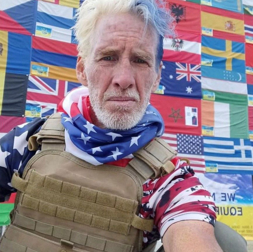 A man in an american flag shirt and army coloured vest takes a selfie 