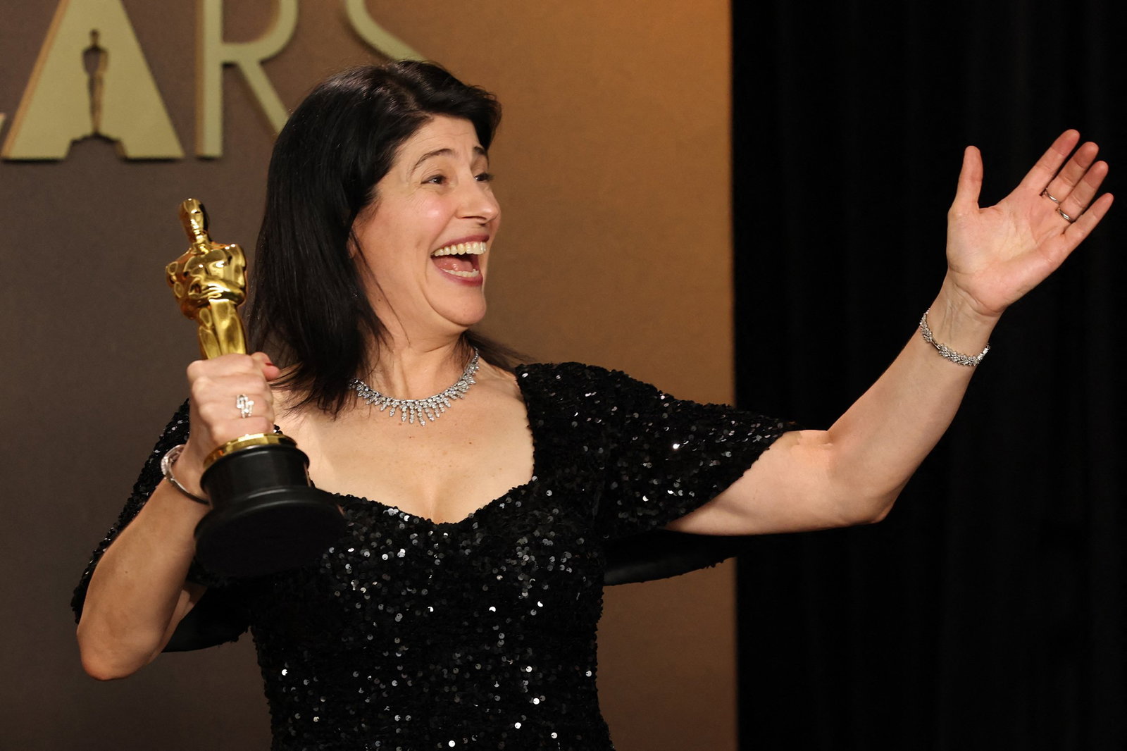 A woman in a black sequinned dress laughs as she holds her Oscar in her right hand and holds her left arm up.