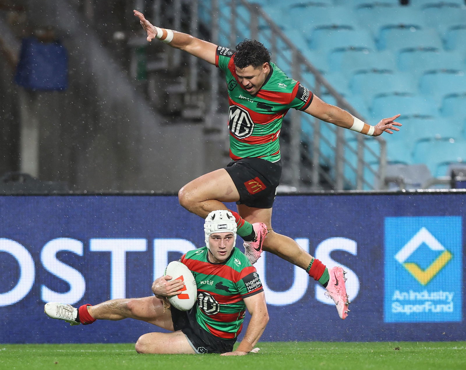 A player holds the ball as he slides on the ground over the try line and a teammate jumps over the top 