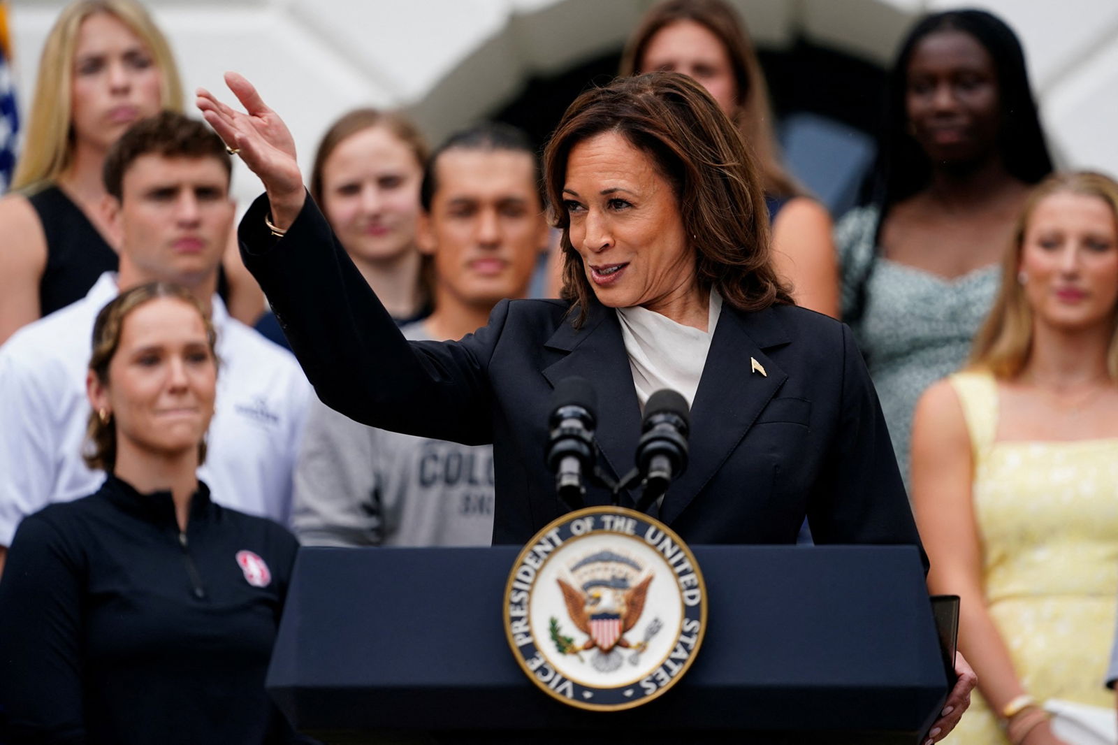 Kamala Harris smiles while raising a hand high, standing at a podium in front of a crowd