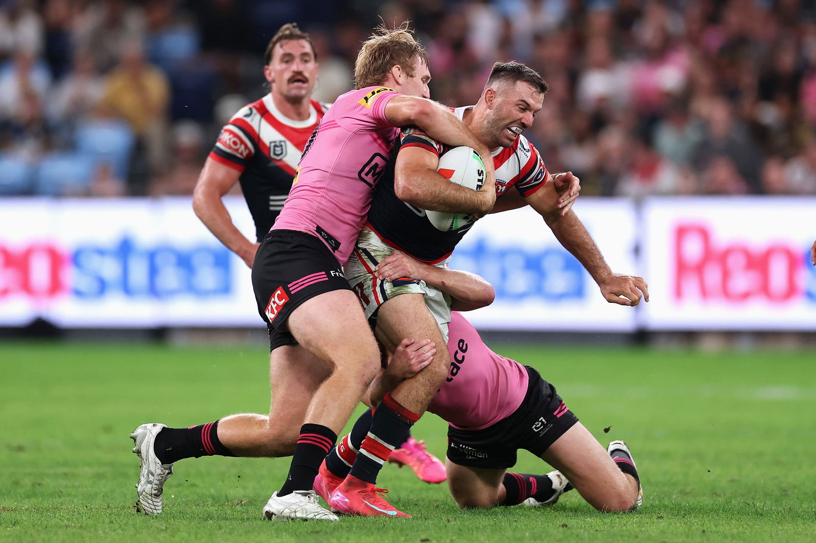 A rugby league player in blue carries the ball while being tackled by two players in pink. Crowd in background.