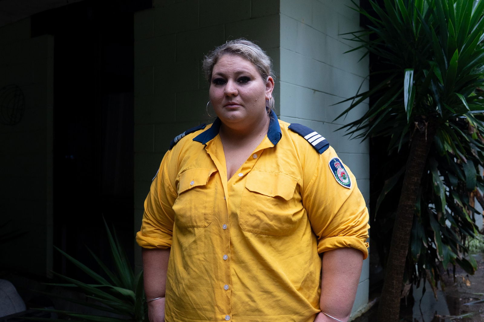 A woman in a yellow shirt with blonde hair stands outside a white brick building