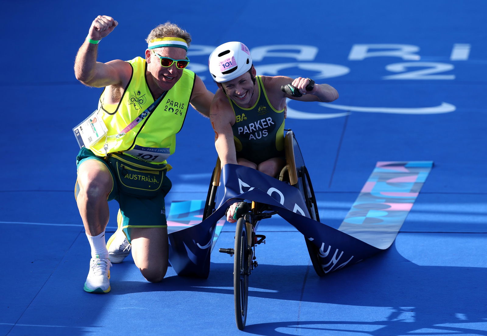 A woman in a racing wheelchair has her fist in the air, next to a man in a fluorescent yellow shirt. Both have an arm in the air in celebration. The woman has a ribbon draped over her and they are on a blue surface.