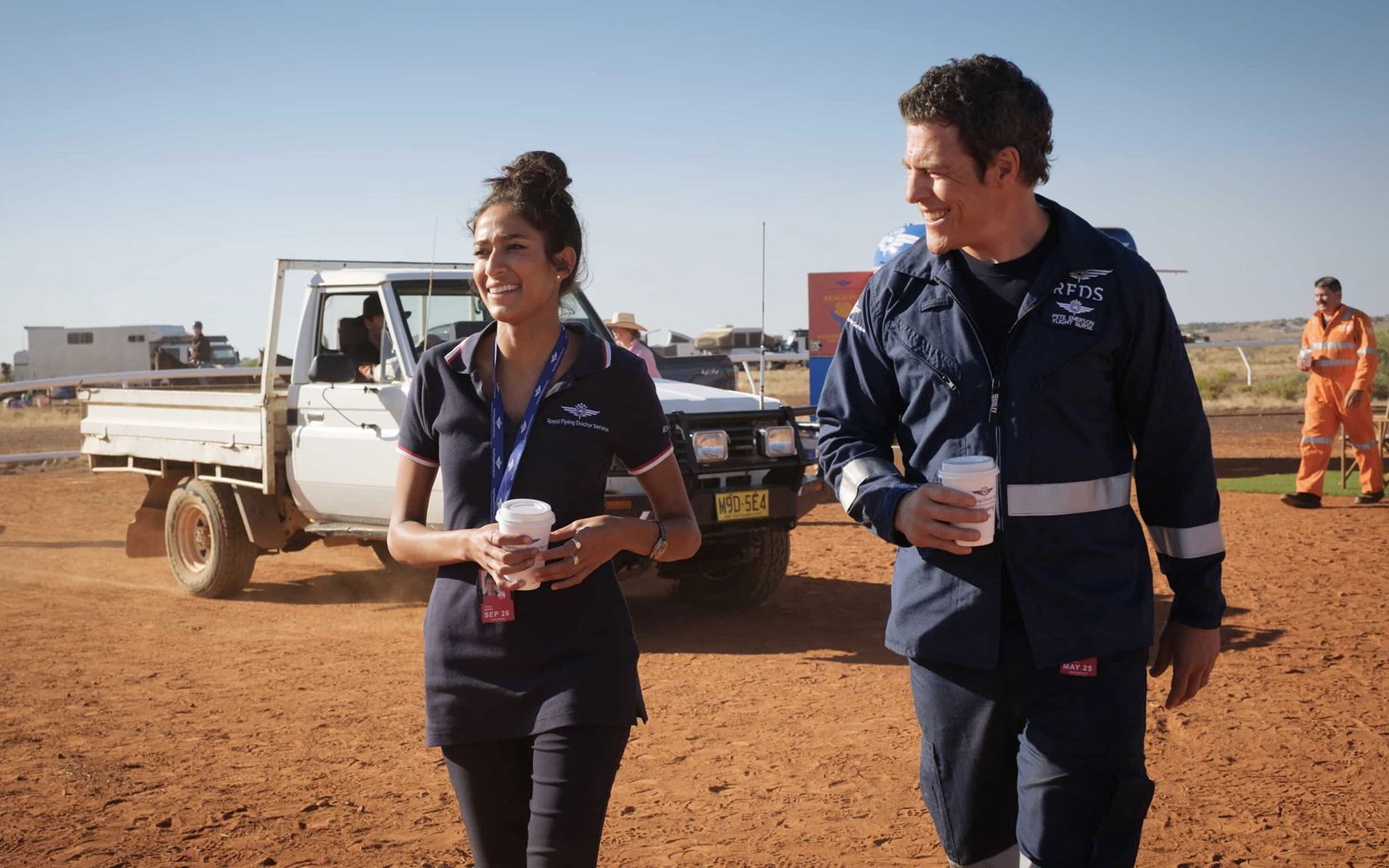 A TV still of Emma Harvie and Stephen Peacocke in RFDS. They're wearing medical uniforms and walking through a bush landscape.