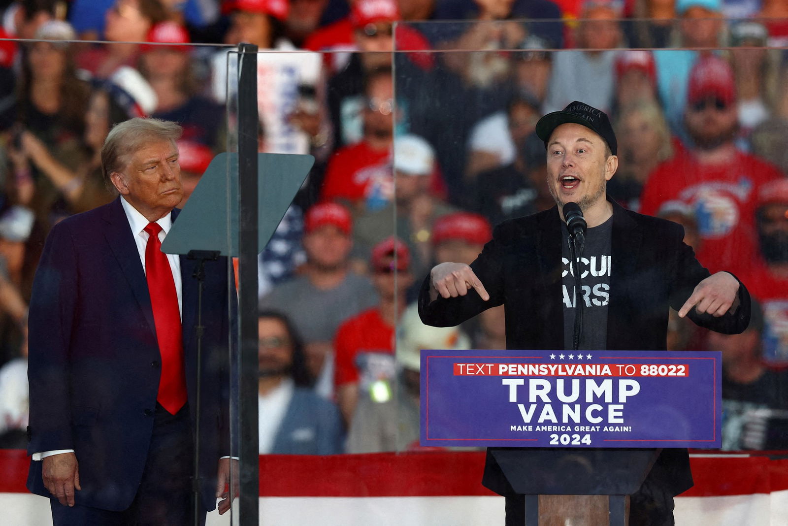 Elon Musk stands at a podium before a crowd. Donald Trump looks on.