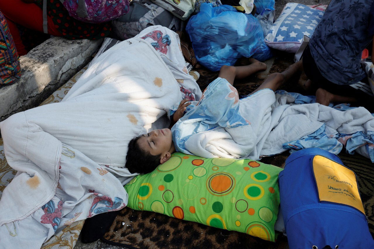 A young boy sleeps on the ground. Other people are visible nearby.