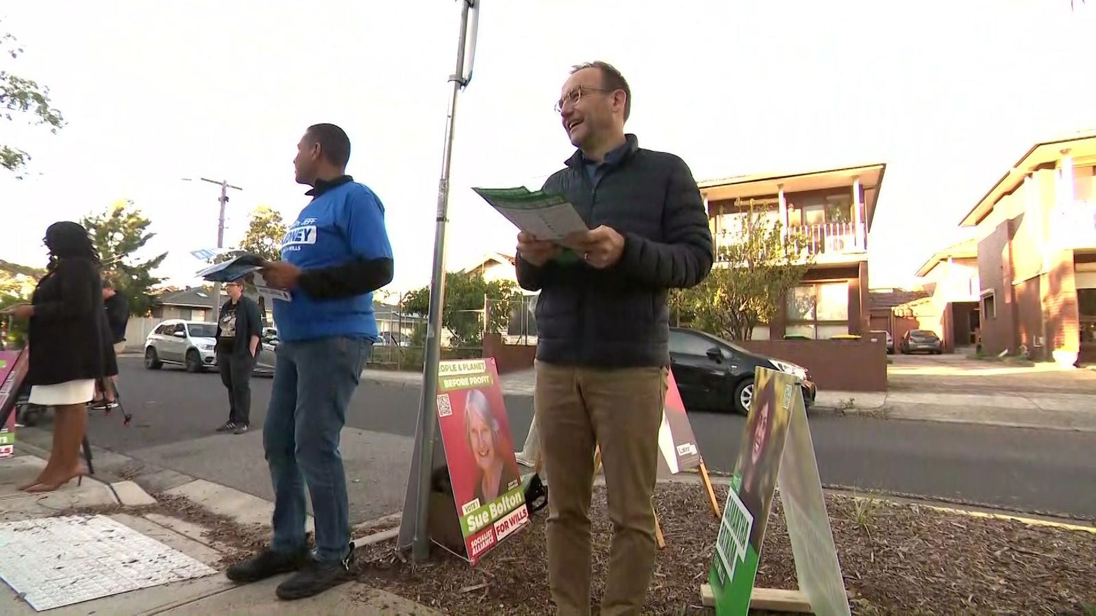 ADam Bandt smiling in a black puffer jacket standing along a road holding how to vote cards