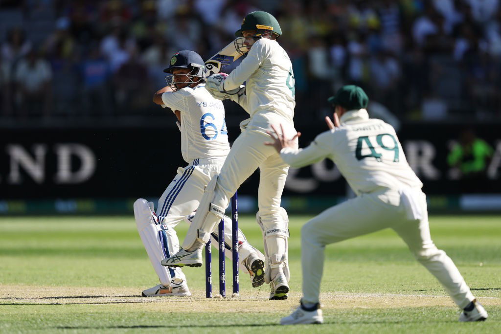 Indian batsman Yashasvi Jaiswal, seen from behind, cuts away as Australian fielders Alex Carey and Steve Smith try to field the ball.