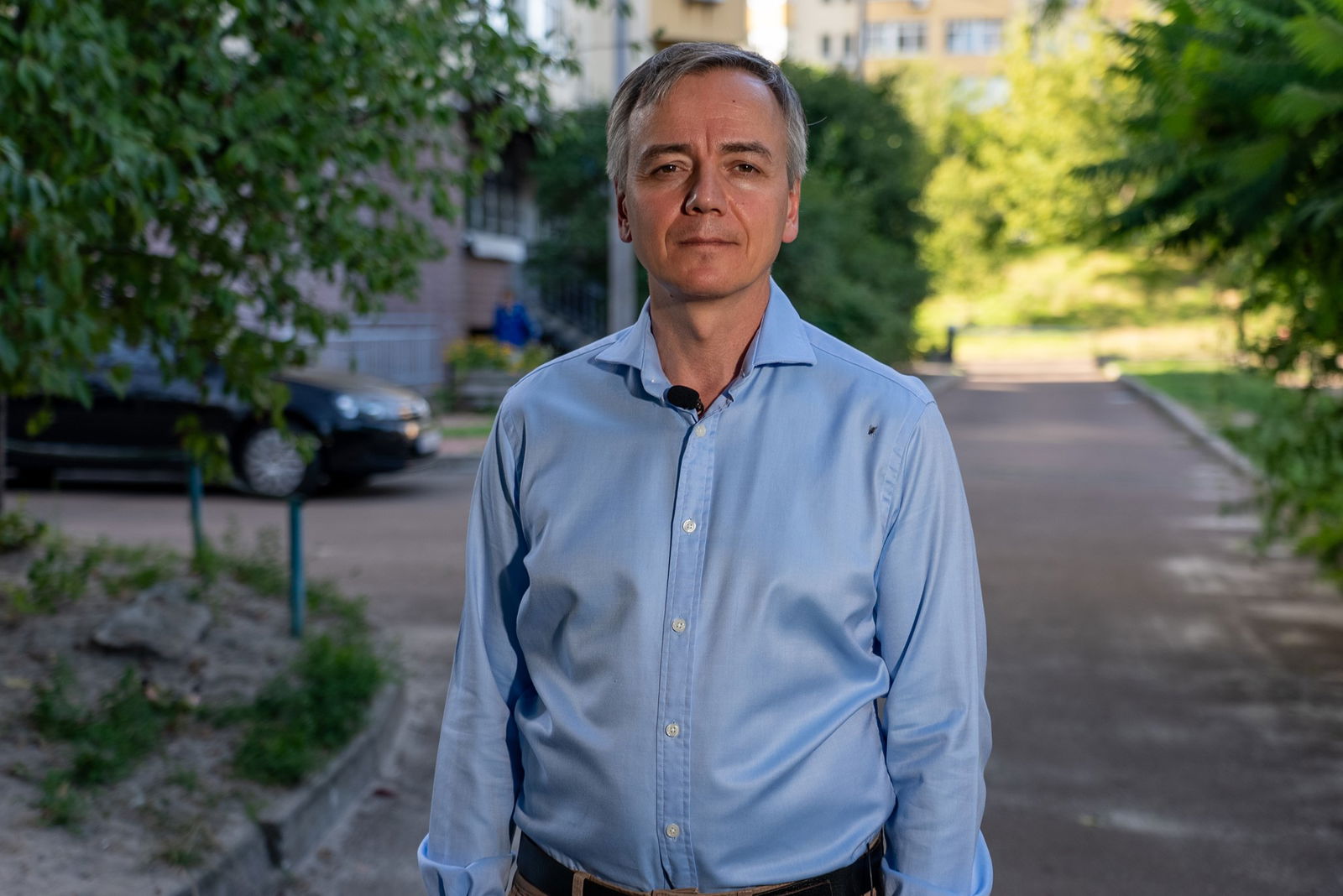 A man in a blue shirt stands looking neutrally at the camera on a street lined with trees.