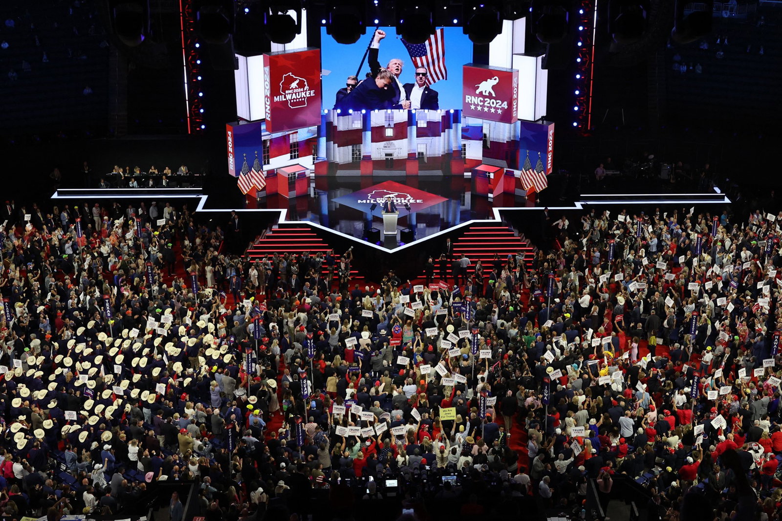 A shot of a large crowd inside a stadium looking to a stage with a photo of Donald Trump raising a fist