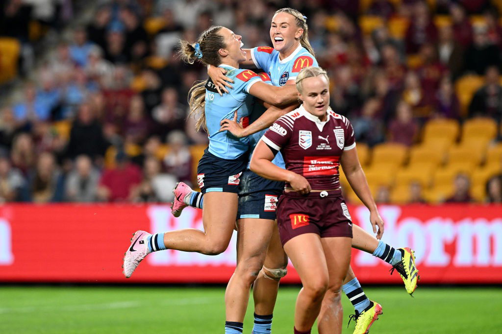 NSW Sky Blues players celebrate a Women's State of Origin try as Queensland's EMily Bass laments in the foreground.