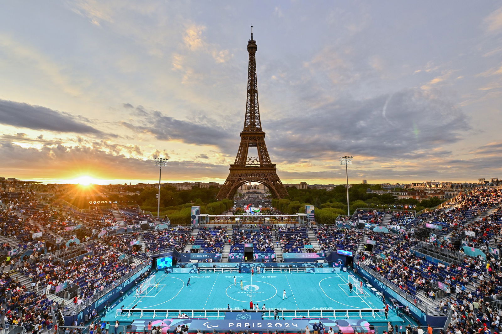 The sun is setting next to the Eiffell tower. The sky is blue and grey and orange. There is a blue blind football court in the foreground, flanked by stands full of spectators.