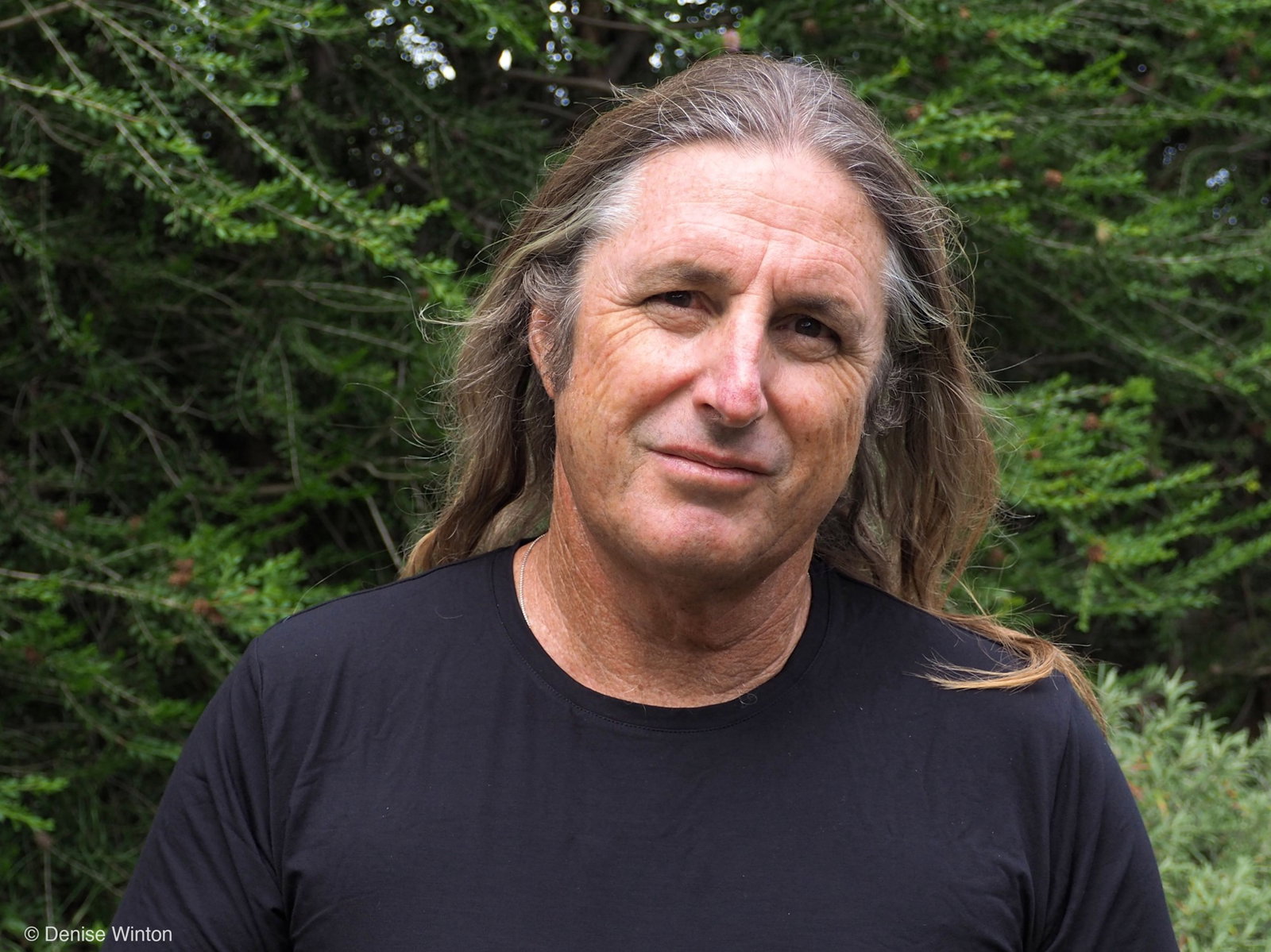 A white man in his 60s with greying long hair, wearing a black t-shirt, standing in front of greenery.