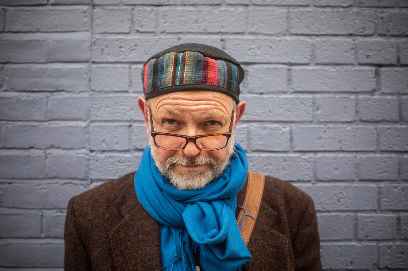 A white man in a brown wool coat, bright blue scarf and multicoloured fabric hat peers over his glasses.