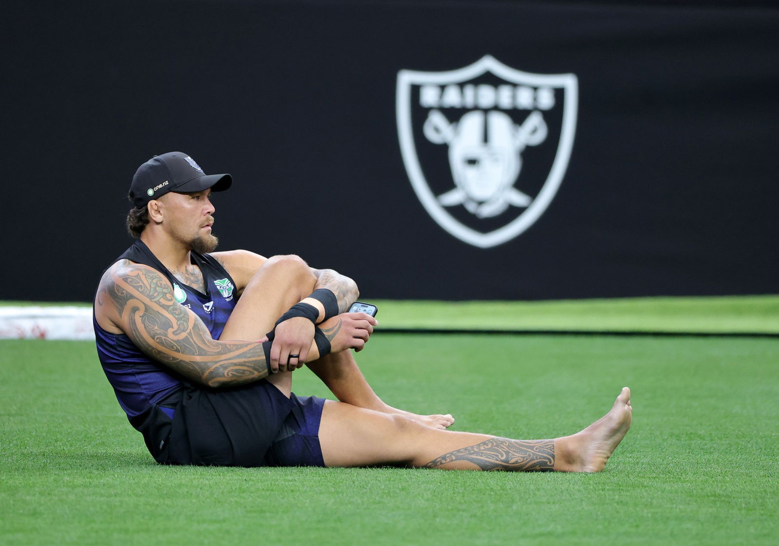 James Fisher-Harris stretches while sitting on the ground with the Las Vegas Raiders logo behind him.