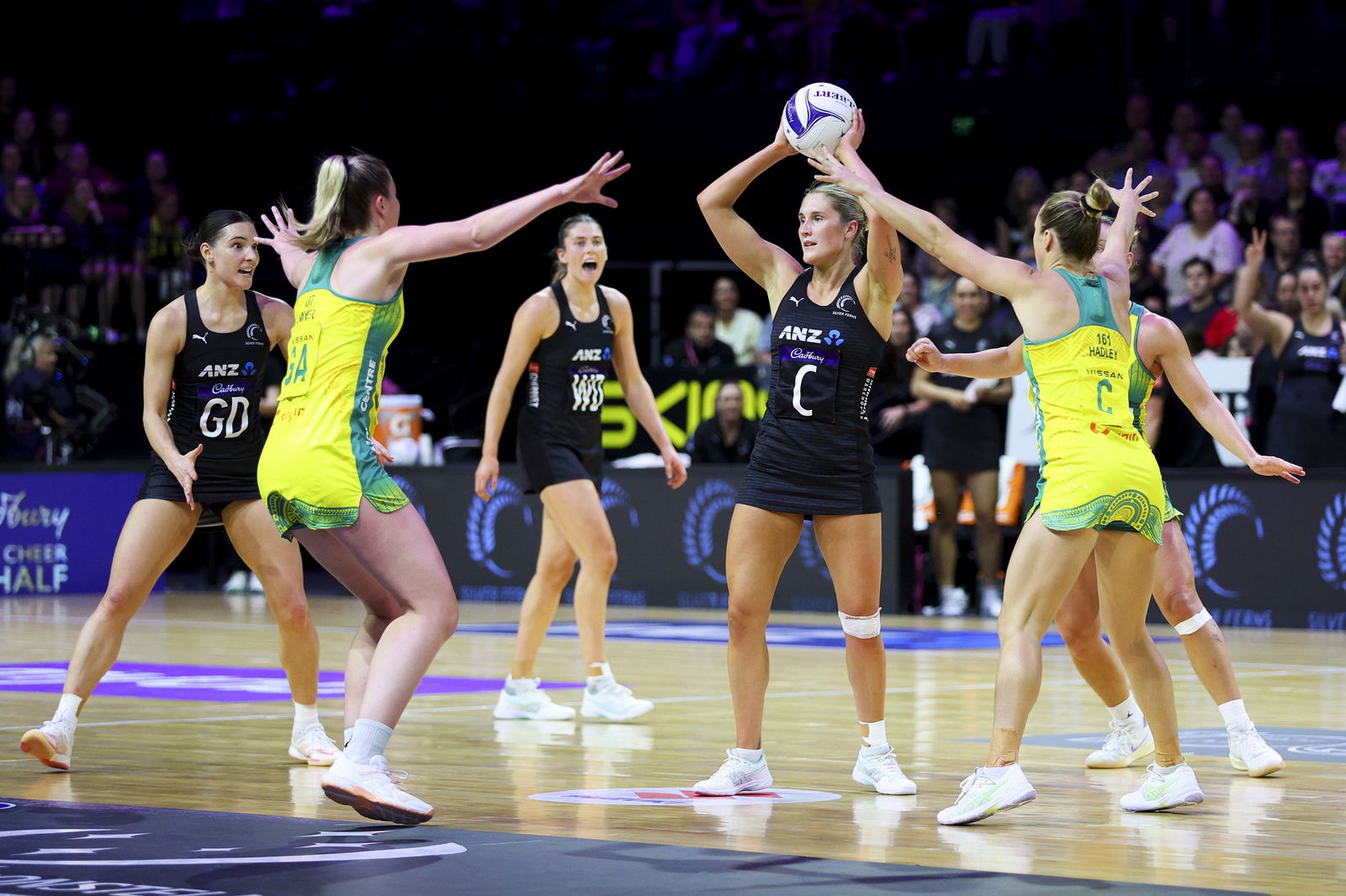 A New Zealand player holds the ball above her head as she is marked by two Australian players