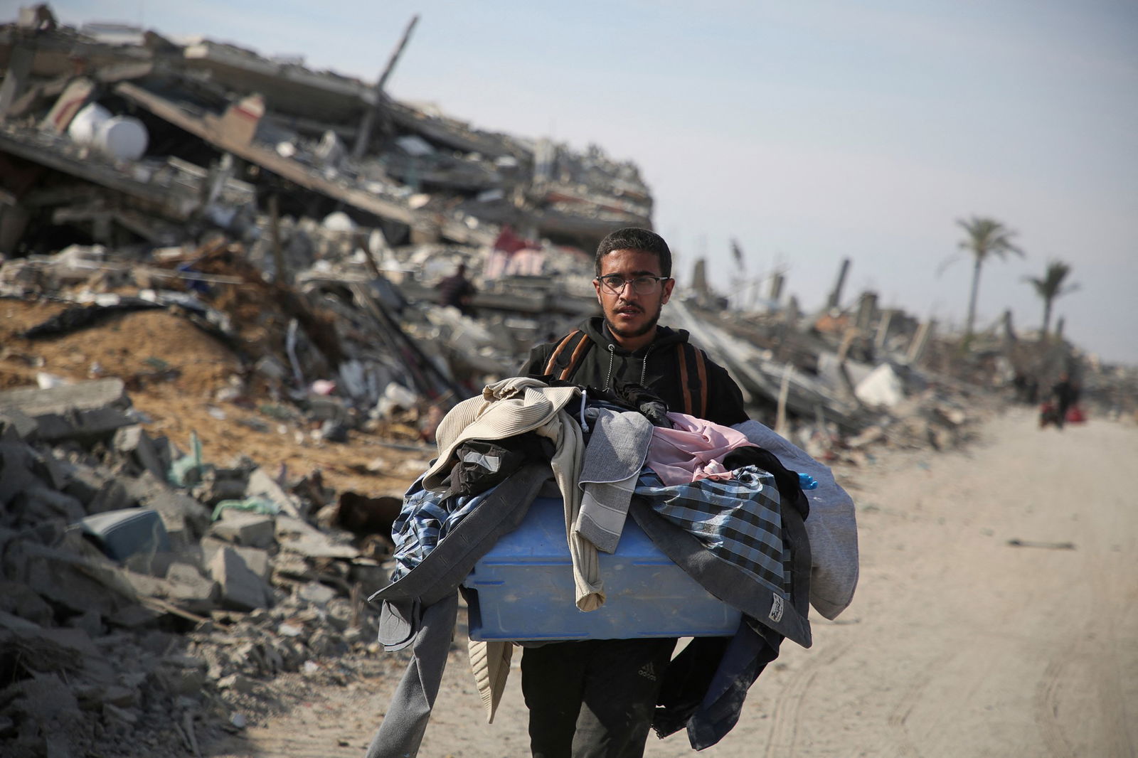 A man carrying a plastic tub full of clothes down a bombed out street.