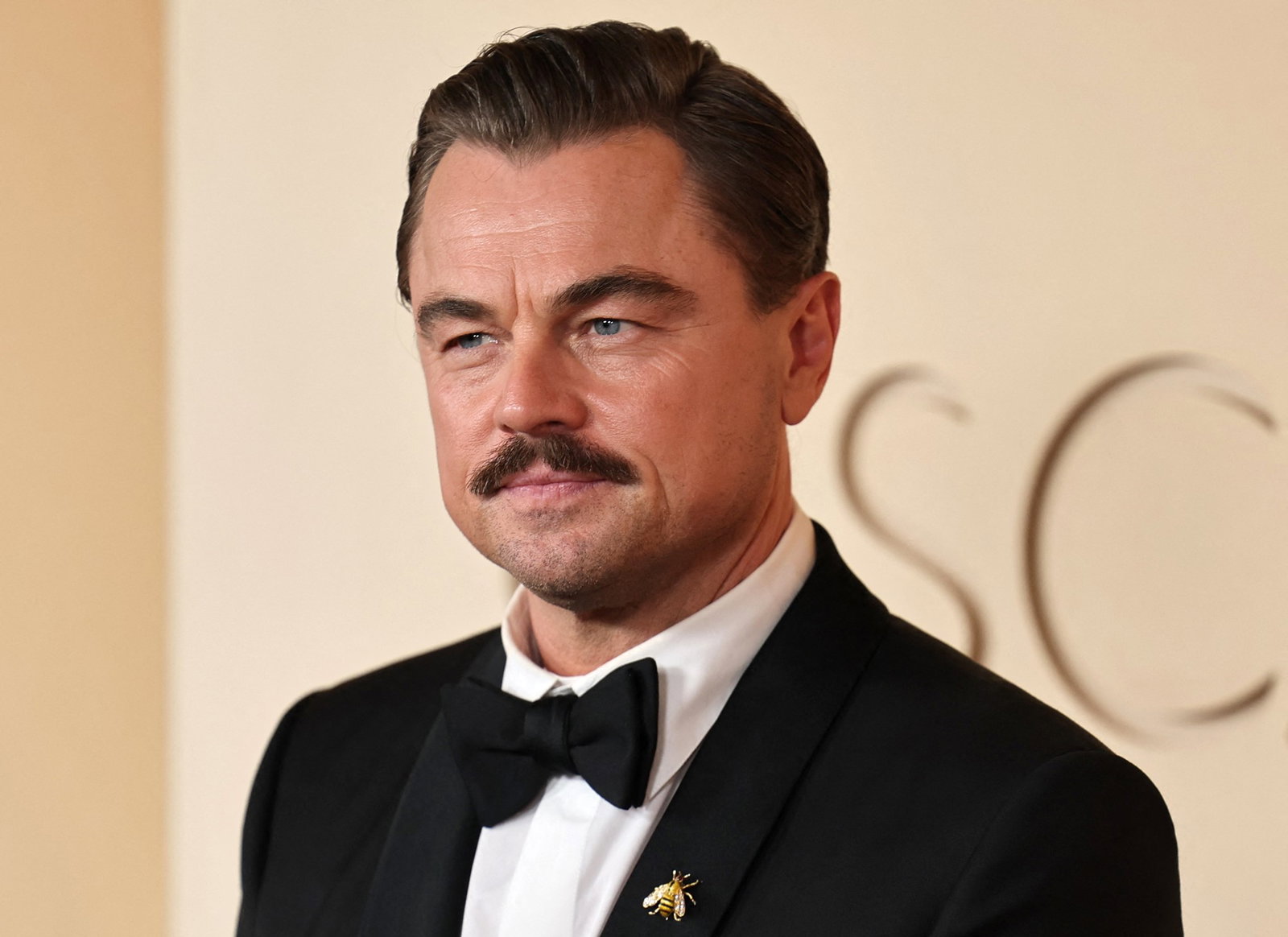 A man with short dark hair and moustache, poses on the red carpet in black suit and tie.
