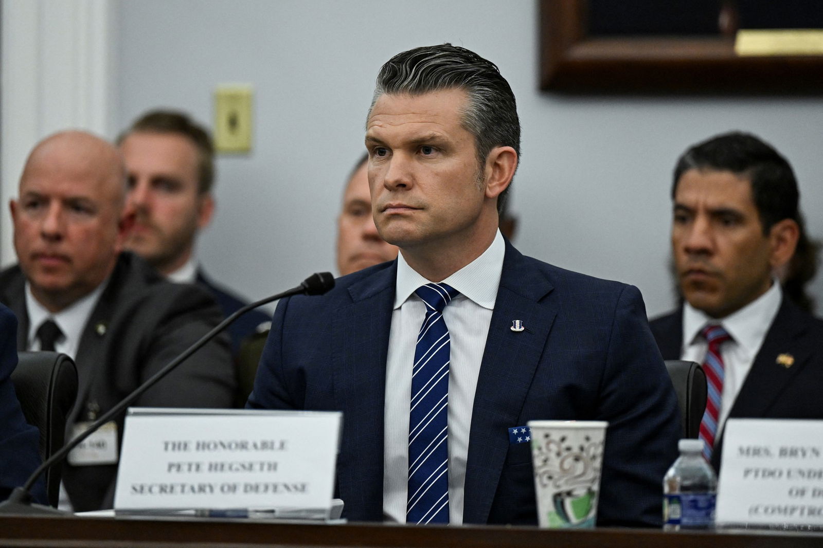 Pete Hegseth sits at a desk with officials behind him
