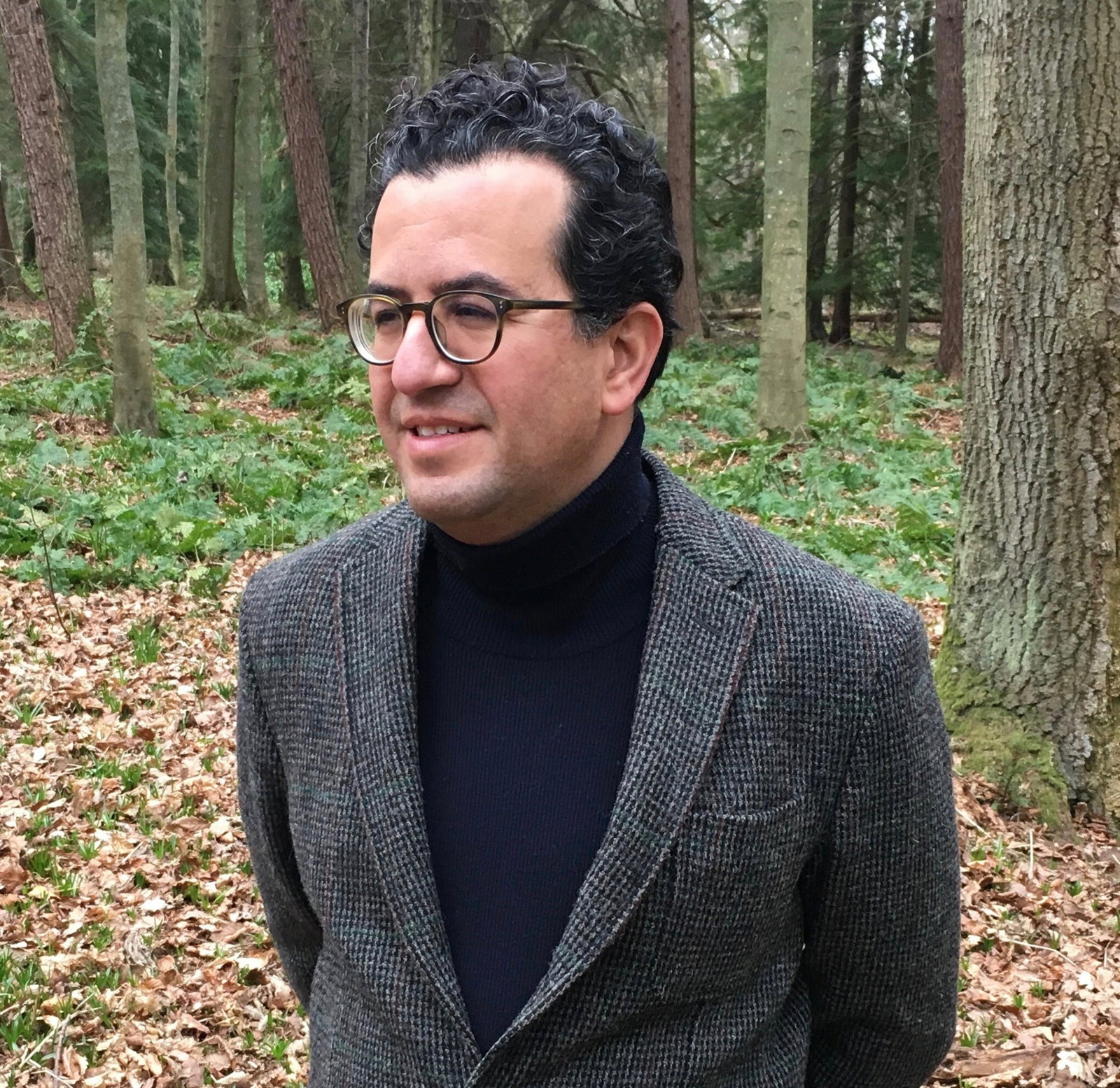 A man with curly black hair and round glasses wears a black turtleneck and tweed jacket, and stands in a forest, looking away.