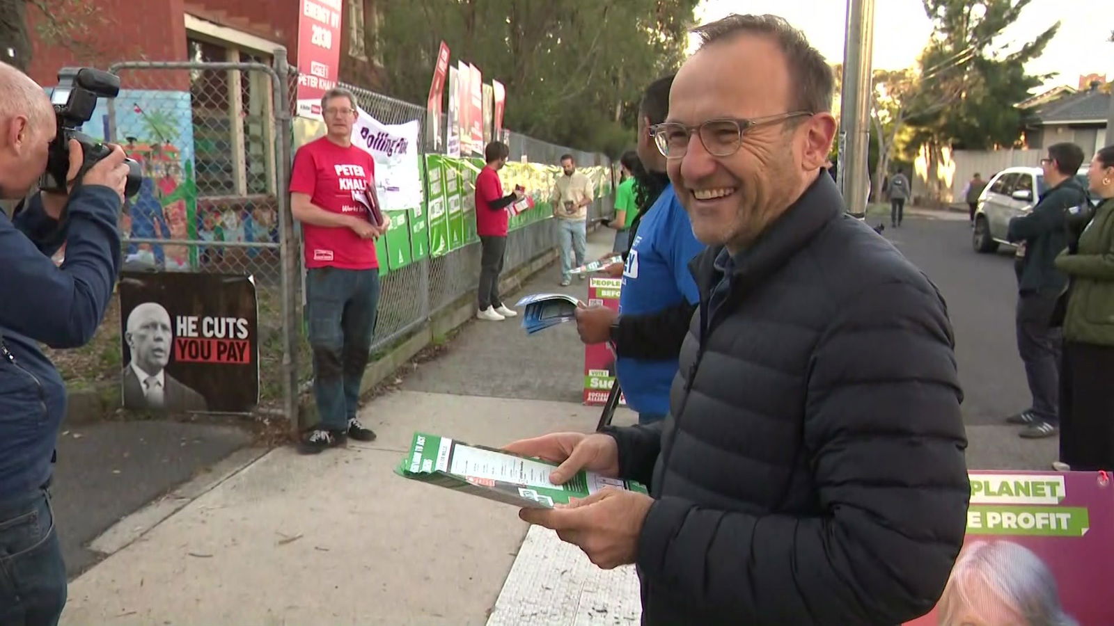 Adam Bandt in a black puffer jacket smiling while holding how to vote cards
