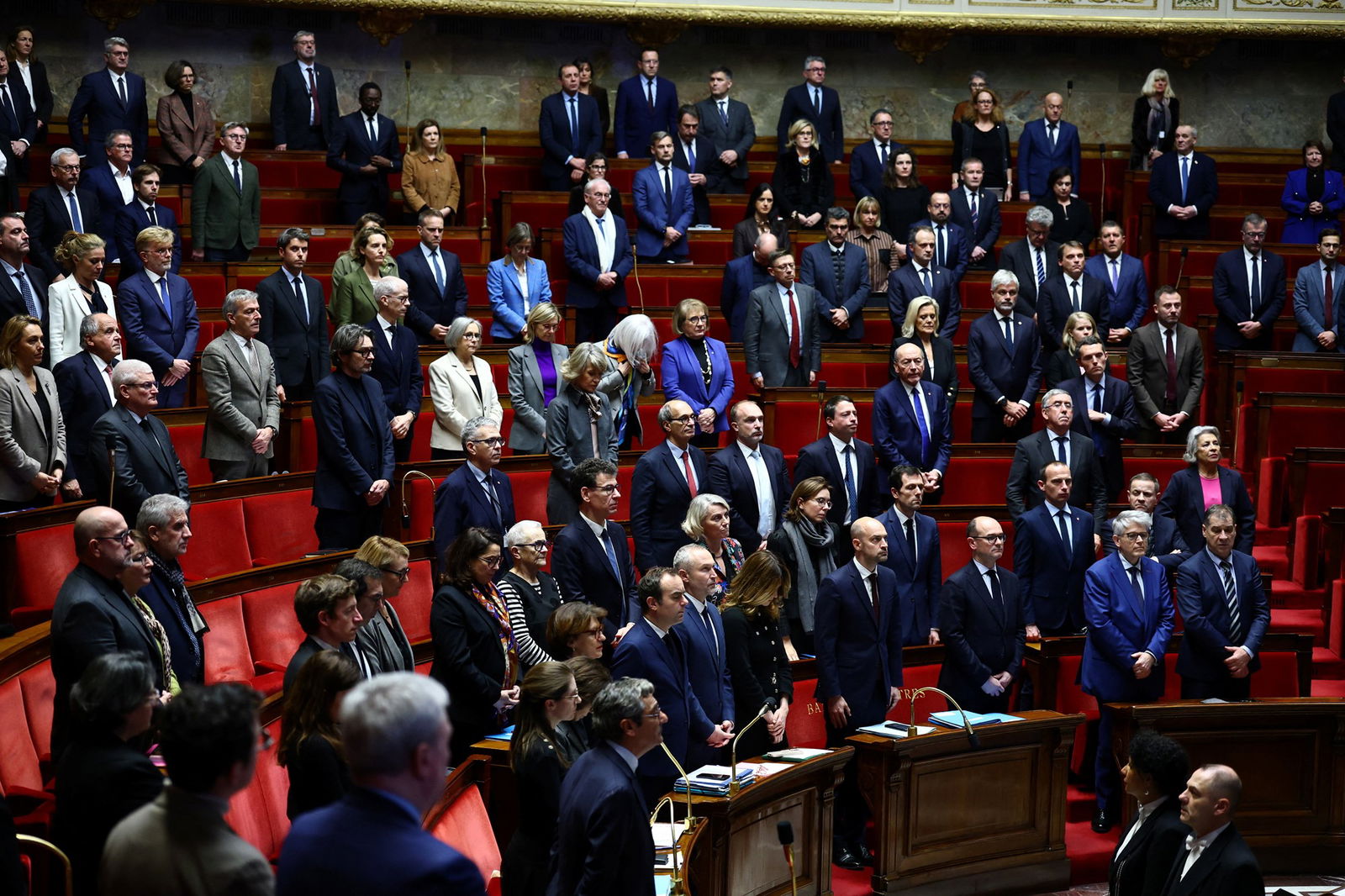 Dezenas de pessoas ficam em uma sala redonda vermelha parecendo sombrias durante um minuto de silêncio.