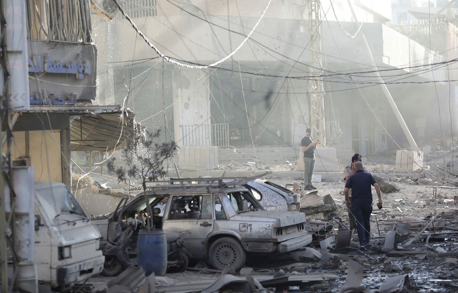 Three people stand amid rubble, including destroyed vehicles