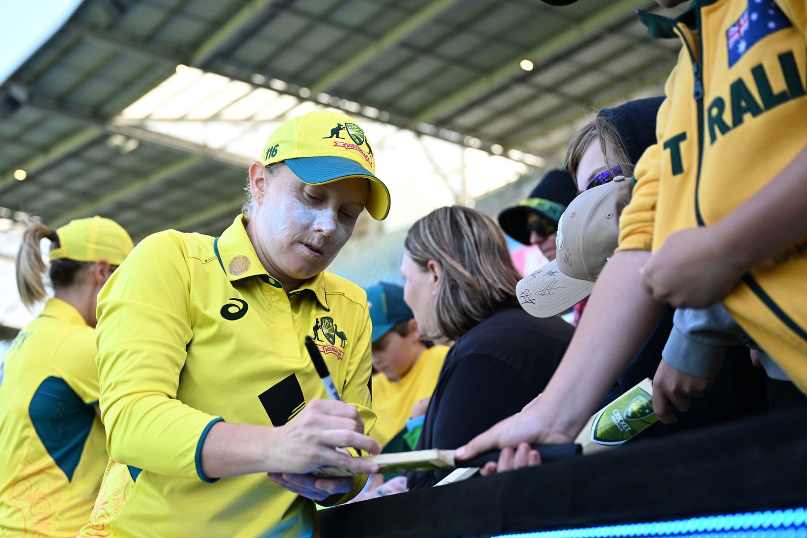 An Australian cricketer in yellow clothes and with white zinc on her face signs a replica cricket bat for a fan.