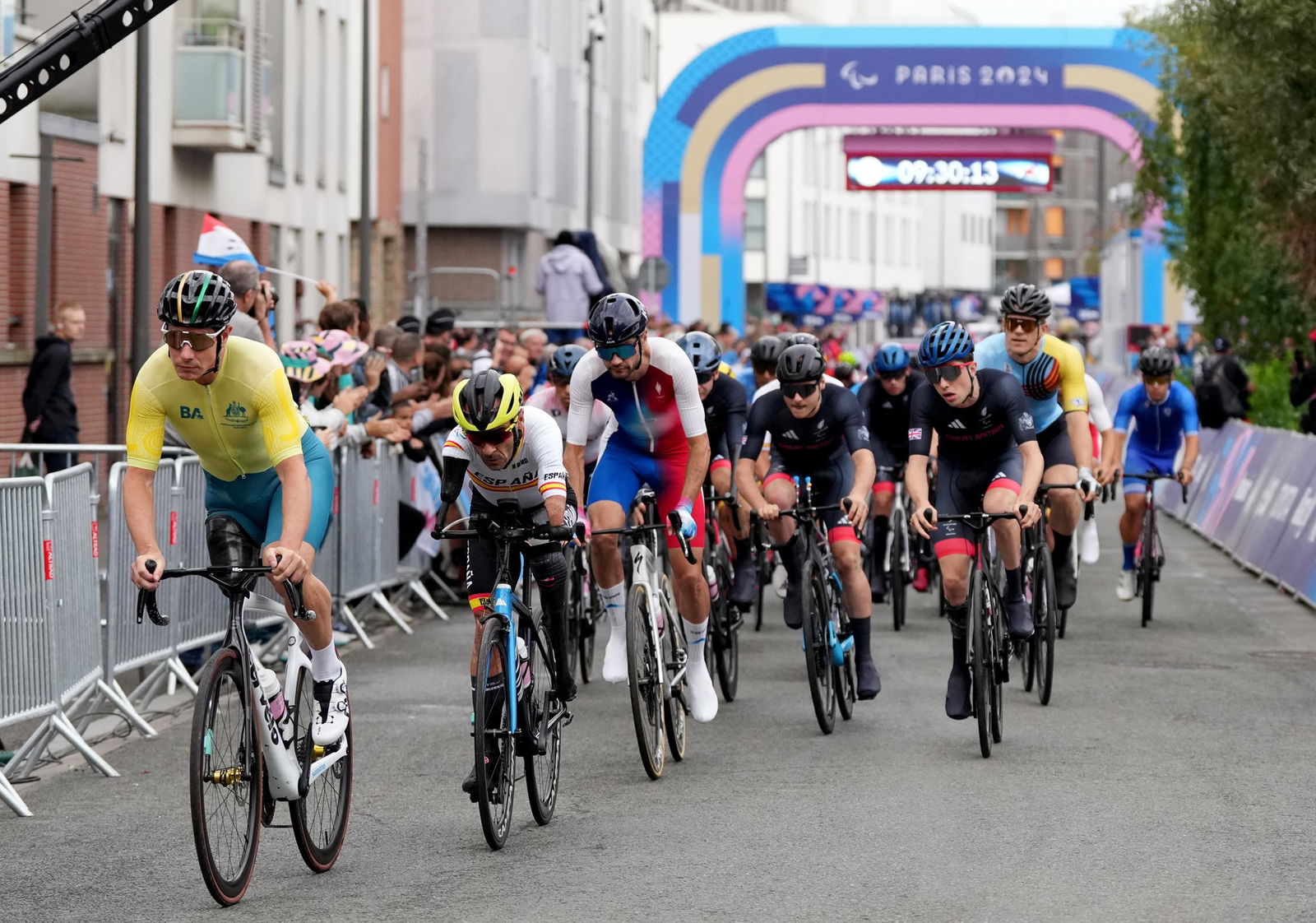 A group of cyclists start a road race, they are all wearing helmets and brightly coloured lycra representing their national colours. The Australian rider is in front wearing green and gold.