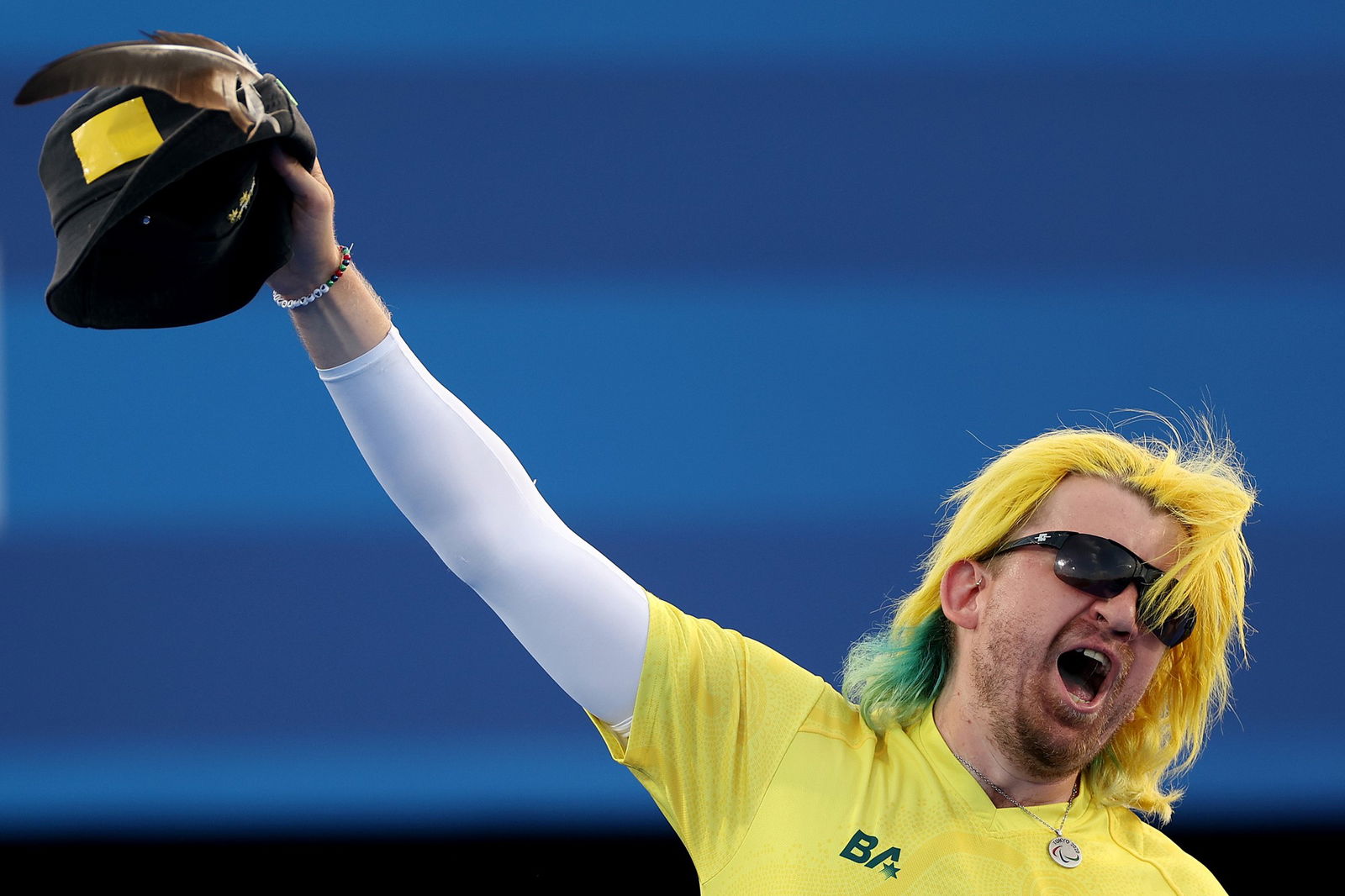 A man with yellow and green hair holds up a hat with a feathern in it. He is wearing sunglasses and shouting in celebration. He is wearing a yellow short sleeved t-shirt with a white undershirt covering his outstretched arm. There is a blue background.