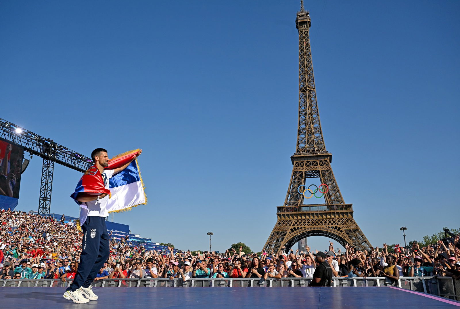 Novak Djokovic drapes a Serbian flag over himself as he shows off his tennis gold medal at the Paris Olympics in front of a crowd at the Eiffel Tower.